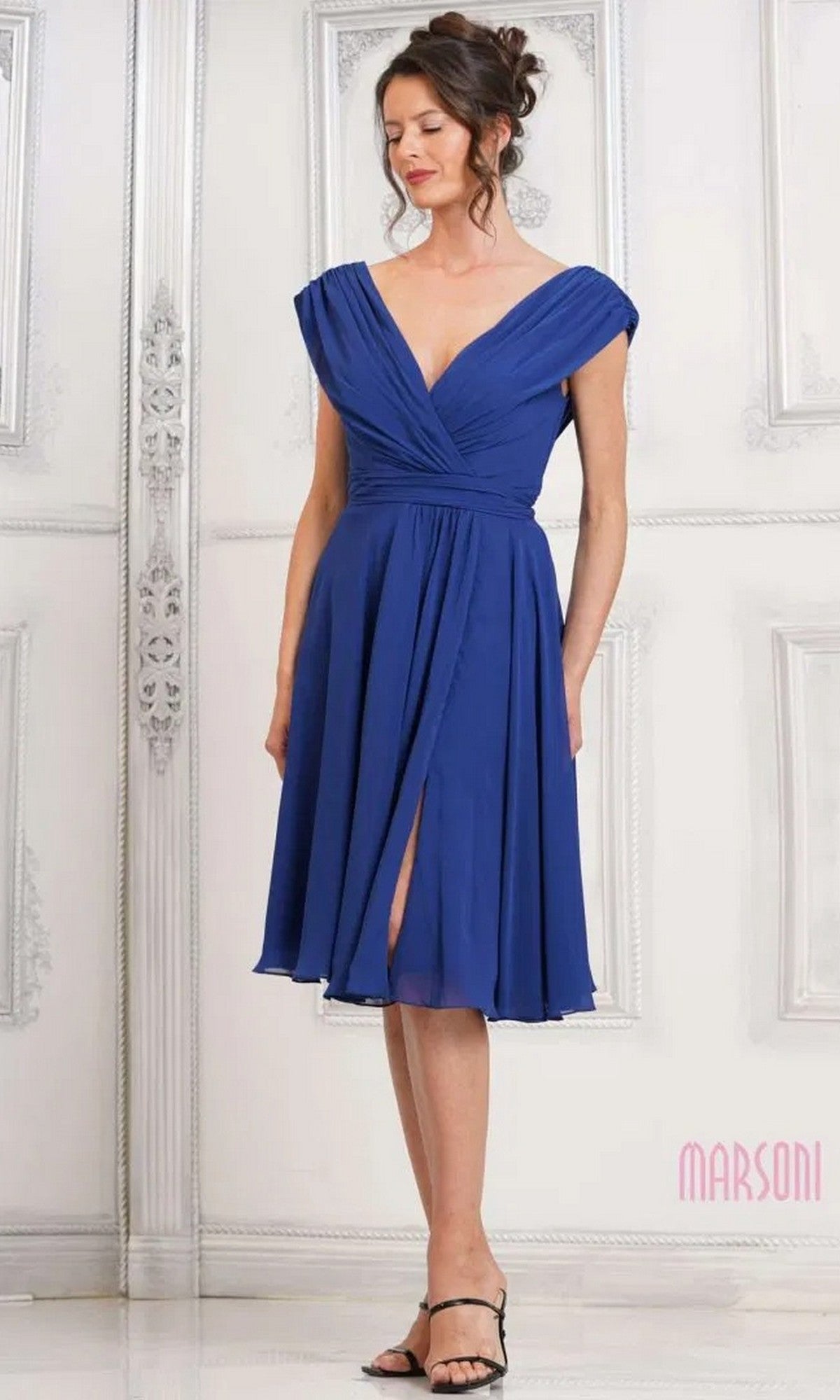 A woman in an indigo blue Marsoni M251S short semi-formal dress with an A-line silhouette and V-neckline stands before a white paneled wall.